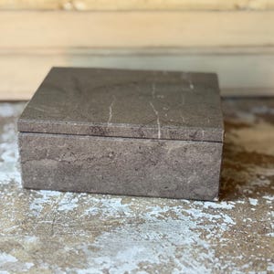 May include: A rectangular box crafted from dark brown stone with natural light-colored veining throughout the surface. The box features a flat, smooth lid that sits flush with the base, showcasing the organic texture and earthy tones of the material.