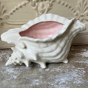 May include: A ceramic conch shell vessel featuring a glossy white exterior with a textured, ridged surface and a smooth, pale pink interior. The shell is shaped with a wide opening and a tapered, pointed end, resting on small, integrated ceramic feet.