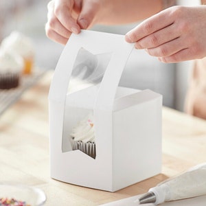 May include: White cardboard cupcake box with a clear window on the front. The box is open and a cupcake is visible inside.