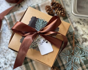 Puede incluir: Una caja de regalo marrón con una cinta de satén marrón atada en un lazo. La caja está decorada con una rama de pino azul y una etiqueta de regalo blanca que dice "To Sara" con un pequeño corazón negro.