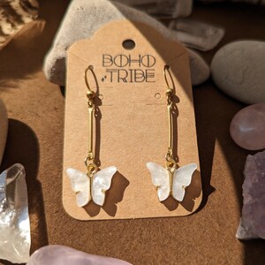 May include: Gold-colored dangle earrings featuring white butterfly pendants. The earrings hang from a gold bar and are displayed on a brown card with the text "BOHO TRIBE". The background includes stones and crystals.