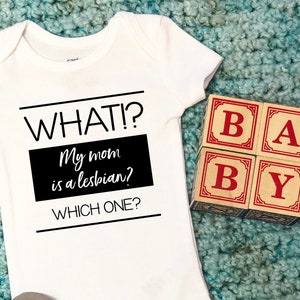 May include: A white baby onesie with the text "WHAT? My mom is a lesbian? WHICH ONE?" in black.  The onesie is on a blue and green carpet with wooden blocks that spell "BABY" in red letters.