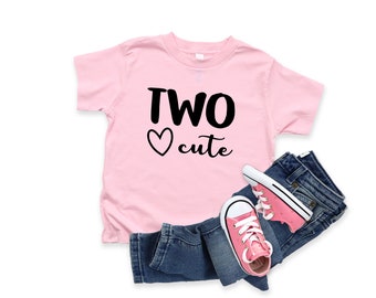 May include: Pink short-sleeved t-shirt with black text that says "TWO heart cute". The shirt is laying on a pair of blue jeans and pink sneakers.