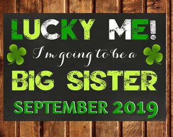 May include: Black chalkboard sign with green shamrock accents announcing "Lucky Me! I'm going to be a Big Sister September 2019."