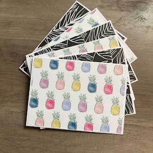 May include: A stack of rectangular cards with white borders. Some cards feature a repeating pattern of colorful watercolor pineapples on a white background. Other cards have a black and white tropical leaf design. The cards are fanned out on a wooden surface.