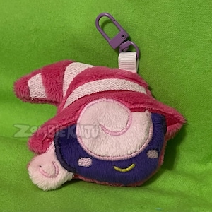 May include: A pink and white striped plush keychain with a purple face and yellow eyes. The keychain has a purple loop for attaching to a bag or other item.
