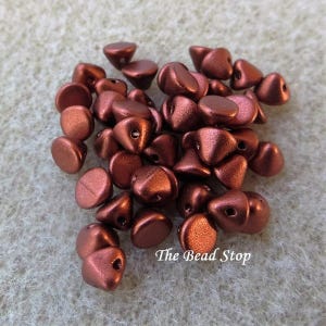 May include: A pile of metallic copper-colored beads. The beads have a unique shape, with a flat base and a rounded, triangular top. Each bead has a small hole for stringing. The text "The Bead Stop" is visible.