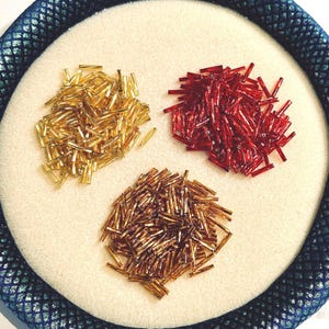 May include: Three piles of small, cylindrical beads in gold, red, and bronze colors. The beads are arranged on a white, circular surface, framed by a dark blue, textured border. These beads are ideal for jewelry making and crafting.