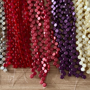 May include: A collection of colorful beads in various shades, including silver, red, pink, purple, and cream. The beads are hexagonal in shape and strung together, creating a visually appealing display of colors and textures.