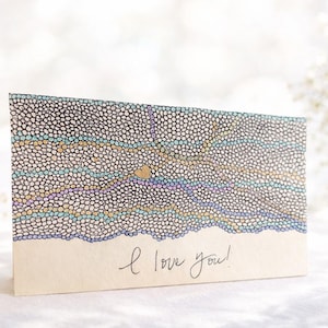 May include: A greeting card with a dotted pattern in shades of blue, purple, and gold, with a small gold heart. The words "I love you!" are written in cursive at the bottom. The card is on a white surface.