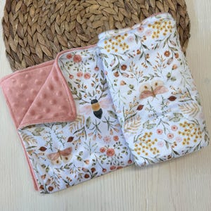 May include: Two baby blankets with floral and insect designs. One blanket has a pink, textured backing. The other two blankets have a white background with a pattern of flowers, leaves, and insects in shades of pink, yellow, and brown.