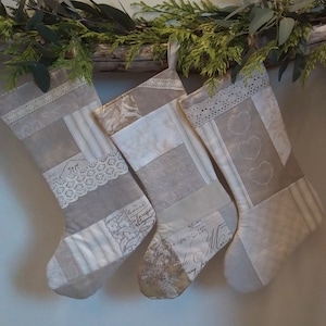 May include: Three patchwork Christmas stockings in neutral beige and cream tones. Each stocking features a mix of fabric patterns, including stripes, floral prints, and script text, accented with white lace trim and embroidered heart details.