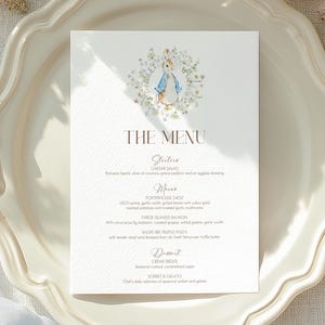 May include: A formal table setting featuring a white plate, gold utensils, and a menu card. The menu card has a floral illustration of a rabbit and the words "The Menu". Soft blue fabric and white flowers surround the setting.