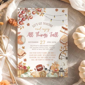Charming autumn harvest invitation with watercolor apples, pumpkins, lantern and fall leaves framing the phrase All Things Fall in warm seasonal colors. Elegant lettering and soft rustic tones give the design a cozy neighborhood gathering feel. The invitation is styled on a round woven placemat surrounded by small white pumpkins, dried florals and soft neutral fabrics that enhance the fall harvest theme.