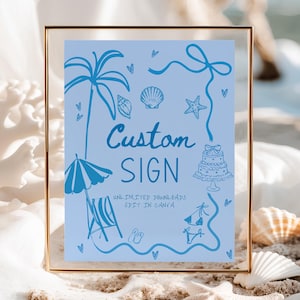 May include: A framed sign with a light blue background and blue illustrations of beach elements, including a palm tree, seashells, and a cake. The words "Custom SIGN" are written in a cursive font. The frame is gold.