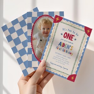 Holding a Fun To Be One first birthday invitation, a hand presents the colorful card alongside the matching checkerboard photo back. Bright vintage inspired typography and whimsical hand drawn elements frame the party details. The reverse side features a personalized child photo within a decorative oval frame. Natural window light casts soft shadows across the scene for an airy modern look.