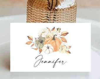 May include: A white rectangular place card featuring a watercolor illustration of pumpkins, white flowers, and autumn leaves. The name Jennifer is printed in black cursive script below the floral arrangement. The card is positioned in front of a textured glass vase.