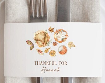 May include: A white rectangular paper place card featuring a watercolor illustration of a roasted turkey, pumpkin, and autumn leaves. The text reads THANKFUL FOR Hannah in brown lettering. The card is positioned over a beige napkin with a fork and knife.