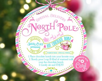 Könnte beinhalten: Ein runder, rosa-weißer Weihnachtsschmuck mit den Worten "North Pole" und "Hot Chocolate Bomb". Der Schmuck hat einen rosa-weißen gestreiften Rand und enthält Gebrauchsanweisungen. Der Text "Santa Claus Approved" ist ebenfalls sichtbar.