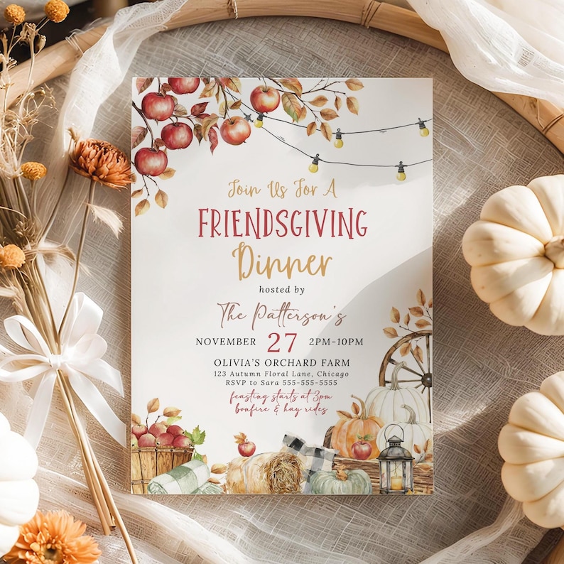 Warm Friendsgiving Invitation displayed upright among white pumpkins and woven textures for a charming harvest presentation. Watercolor apples, pumpkins, lanterns and hay bales frame the invitation while delicate string lights add a cozy touch. The surrounding autumn decor creates an inviting Thanksgiving setting. This image emphasizes the rustic fall dinner party theme and seasonal artwork.