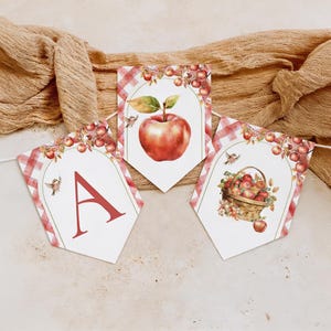 May include: A decorative bunting banner featuring three pennants with a red and white checkered border. The pennants display a red letter A, a large red apple, and a woven basket filled with red apples, all accented with small birds and branches.