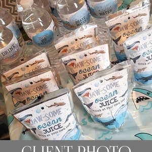 May include: A table with a blue tablecloth and a variety of party favours. The favours are in clear plastic bags with a shark design and the text "JAW-SOME ocean JUICE drink at your own risk". There are also water bottles with labels that say "Fin-Sastic Shark Water" and "JAW-SOME ocean WATER".