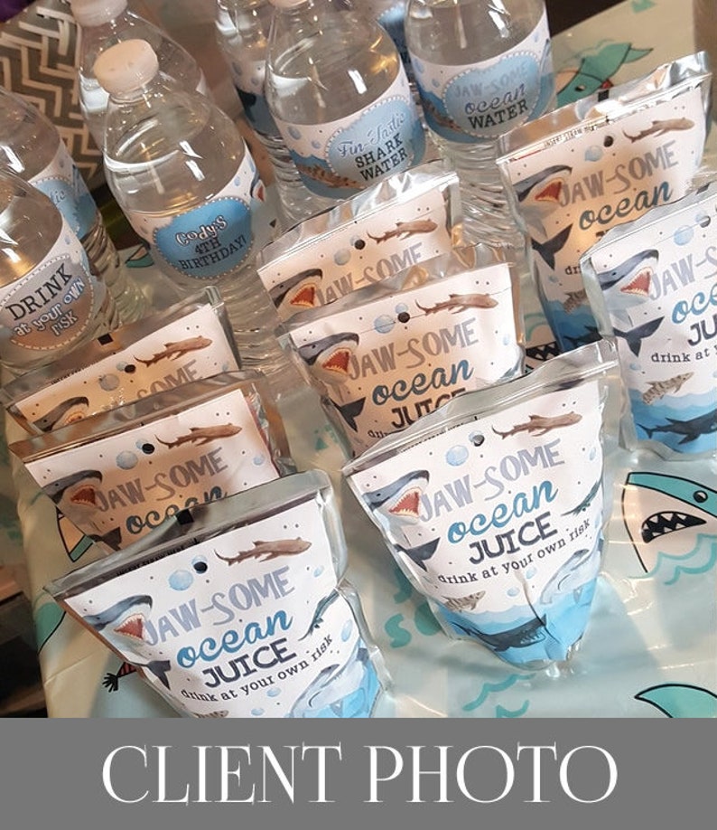 May include: A table with a blue tablecloth and a variety of party favours. The favours are in clear plastic bags with a shark design and the text "JAW-SOME ocean JUICE drink at your own risk". There are also water bottles with labels that say "Fin-Sastic Shark Water" and "JAW-SOME ocean WATER".