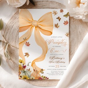 Pastel pumpkin first birthday invitation for girls featuring a large orange coquette bow, watercolor pumpkins, autumn leaves, and butterflies, displayed on neutral linen fabric with soft white peony flowers. Perfect for a chic fall birthday party, rustic pumpkin patch celebration, or pastel autumn gathering with editable and printable personalized text fields.
