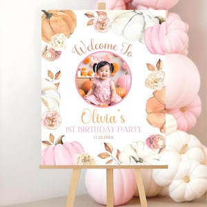 Custom photo welcome sign for a pumpkin first birthday party featuring watercolor pink white and orange pumpkins roses and fall leaves with personalized text and a circular photo of the birthday child displayed on an easel with pastel pink balloons for a charming autumn themed first birthday party backdrop