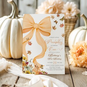 Elegant pastel pumpkin birthday invitation with a large soft orange coquette bow, watercolor pumpkins, leaves, and fall florals, styled upright on a rustic wood surface with cozy seasonal details, including cream pumpkins and soft floral accents. Perfect for a chic autumn birthday party or first birthday pumpkin patch gathering with editable text fields.