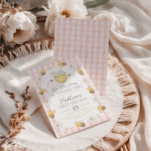 The Printable Occasions offers custom wedding birthday and baby shower templates for every celebration. Editable pink bee birthday invitation with watercolor bumblebees, white daisies, honey jar, and pink gingham border. Displayed on woven placemat with white fabric, dried flowers, and matching gingham back design. Canva template perfect for a girl’s first bee day party.