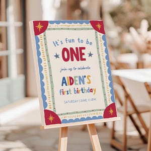 May include: A rectangular birthday sign on a wooden easel featuring a cream background with a blue scalloped border and red corner accents. The text reads: It's fun to be ONE, join us to celebrate, AIDEN'S first birthday, SATURDAY | JUNE 1 | 2PM.