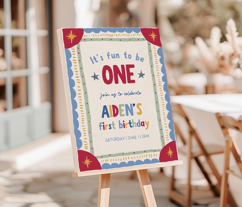 May include: A rectangular birthday sign on a wooden easel featuring a cream background with a blue scalloped border and red corner accents. The text reads: It's fun to be ONE, join us to celebrate, AIDEN'S first birthday, SATURDAY | JUNE 1 | 2PM.