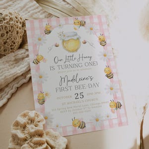 The Printable Occasions offers custom wedding birthday and baby shower templates for every celebration. Pink bee first birthday invitation with watercolor bees, daisies, and honey jar on soft pink checkered border. Shown on neutral textured fabric with coral reef accents and pampas grass. Editable Canva design ideal for a girl’s 1st bee day celebration.