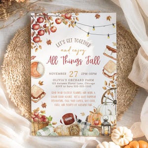 Cozy fall gathering invitation featuring watercolor pumpkins, apples, hay bale, lantern and fall foliage accents arranged around the editable party details. The warm red and golden lettering creates a welcoming seasonal look for a neighborhood fall celebration. The card rests on a natural woven placemat with airy fabric and soft neutral decor creating a light autumn styled scene.