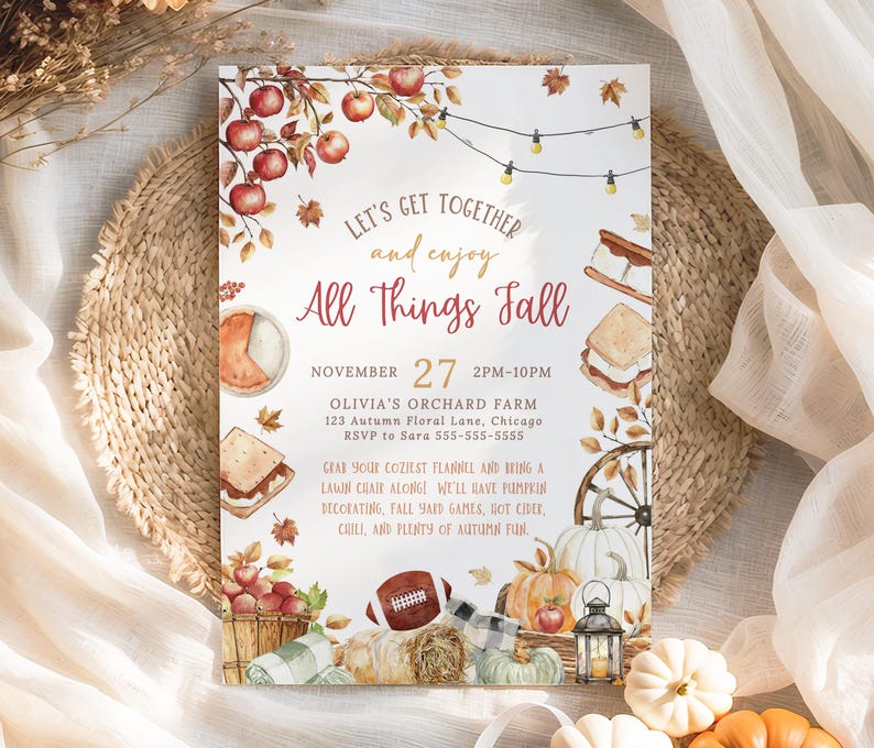Cozy fall gathering invitation featuring watercolor pumpkins, apples, hay bale, lantern and fall foliage accents arranged around the editable party details. The warm red and golden lettering creates a welcoming seasonal look for a neighborhood fall celebration. The card rests on a natural woven placemat with airy fabric and soft neutral decor creating a light autumn styled scene.