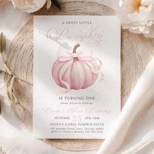 Pink pumpkin first birthday invitation with a large soft watercolor pumpkin and pastel pink ribbon bow in the center, perfect for a fall baby girl birthday. Elegant text reads “A Sweet Little Pumpkin” with custom party details below. The invite is styled flat on a woven neutral placemat with white peonies and light linen fabric, giving the scene a cozy rustic autumn vibe. Soft shadows and sunlight enhance the warm fall aesthetic.