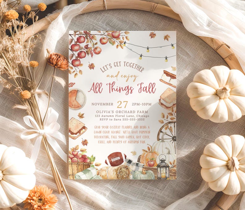 Charming autumn harvest invitation with watercolor apples, pumpkins, lantern and fall leaves framing the phrase All Things Fall in warm seasonal colors. Elegant lettering and soft rustic tones give the design a cozy neighborhood gathering feel. The invitation is styled on a round woven placemat surrounded by small white pumpkins, dried florals and soft neutral fabrics that enhance the fall harvest theme.