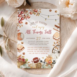 Seasonal fall festival invitation design with watercolor pumpkins, apples, hay bale, lantern and autumn leaves surrounding the party wording in warm harvest colors. The center text area allows customization of date, location and event details. The card is displayed upright with soft pumpkins, dried florals and warm fall tones in the background creating a cozy autumn gathering scene.