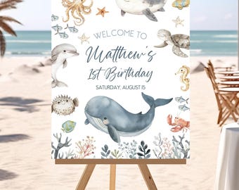 May include: A white welcome sign featuring a blue whale, a dolphin, an octopus, a starfish, a seahorse, a turtle, and a crab. The sign reads "Welcome to Matthew's 1st Birthday, Saturday, August 15".