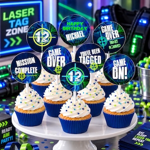 May include: A set of round cupcake toppers featuring laser tag themes with text including "12", "Happy Birthday Michael", "Game Over", "Mission Complete", "You've Been Tagged", and "Game On!" displayed on cupcakes with white frosting and blue liners.
