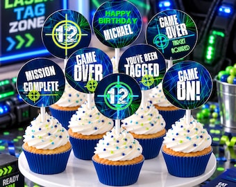 Puede incluir: Un conjunto de siete adornos redondos para cupcakes con temas de laser tag y textos que incluyen Happy Birthday Michael, 12, Game Over, Mission Complete, You've Been Tagged y Game On, sobre cupcakes con glaseado blanco en moldes azules.
