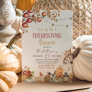 Lovely Friendsgiving invitation shown with the optional orange gingham backside and coordinating harvest inspired front design. The invitation features watercolor apples, pumpkins and string lights that complement the warm orange gingham pattern. The cards are displayed on soft neutral fabric with dried florals and seasonal decor surrounding them. This image showcases the second included backside color option for the printable invitation.