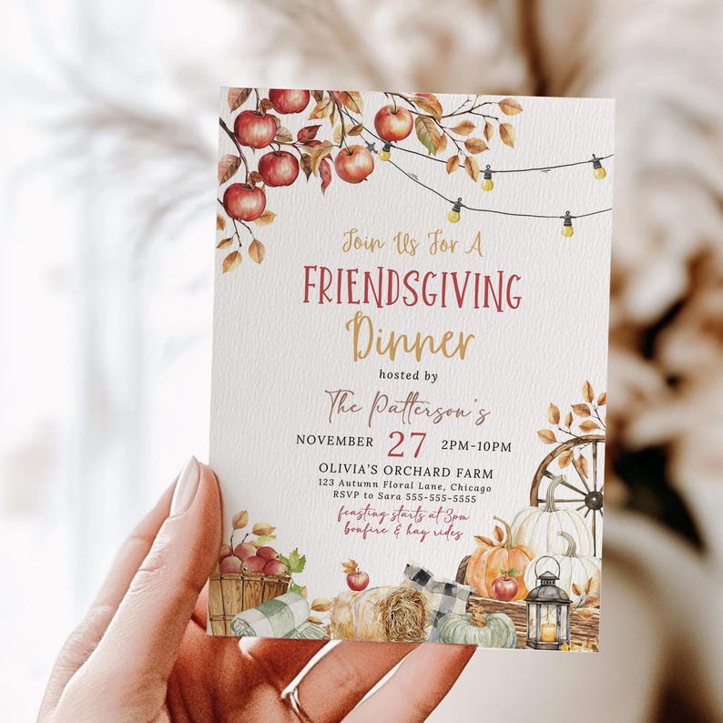 Beautiful Editable Friendsgiving Invitation featuring watercolor apples hanging from branches, string lights, pumpkins, hay bales and rustic harvest details in warm autumn colors. The invitation is held in a hand to showcase the size and printed texture of the design. The softly blurred neutral background creates a cozy and inviting fall atmosphere. This image highlights the rustic fall dinner party invitation and elegant seasonal artwork.