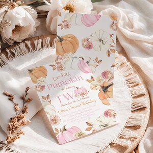 The Printable Occasion offers a girl’s second birthday invitation with a pink pumpkin theme. The design features watercolor pumpkins, blush florals, and autumn leaves with the wording “Our Little Pumpkin is Turning Two.” Editable in Canva, this fall birthday invitation is shown in a flatlay on a rustic woven tray with white fringe linen, flowers, and natural light styling.