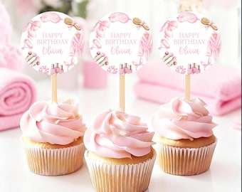 May include: Three cupcakes with pink frosting and decorative toppers. The toppers are round with a spa-themed design and the text "HAPPY BIRTHDAY Olivia". The cupcakes sit on a white surface, with pink towels in the background.