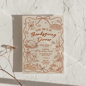 Thanksgiving dinner invitation displayed upright against a textured cream wall with soft natural light and subtle rainbow reflections. The hand drawn design includes a turkey, pumpkin, pie, wine glass, candles, leaves and bow details in warm brown on cream. Delicate dried stems appear along the lower left. The full 5x7 invitation design is clearly visible.