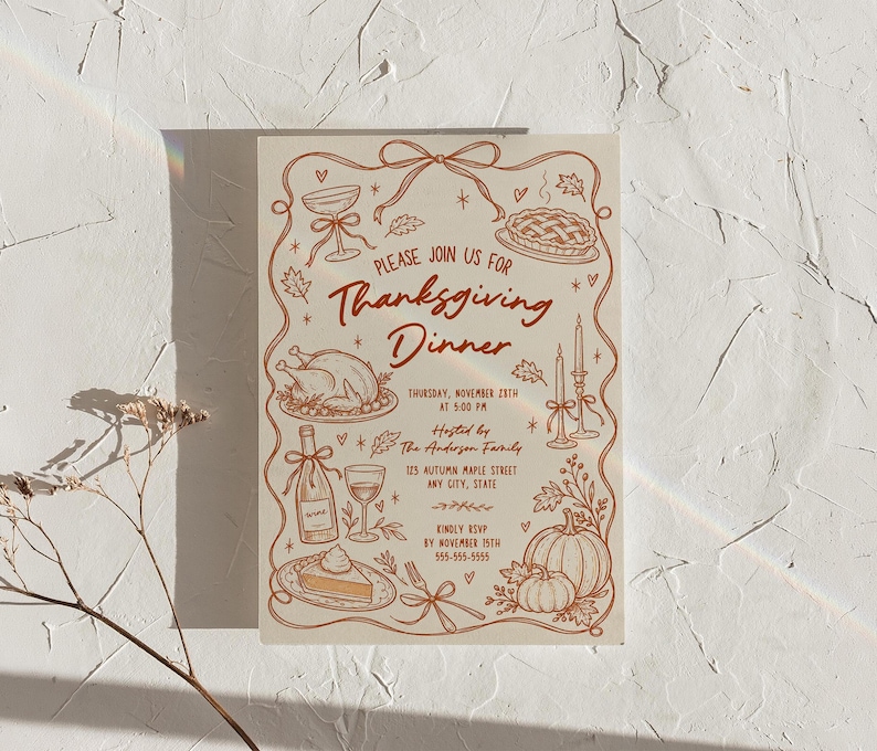 Thanksgiving dinner invitation displayed upright against a textured cream wall with soft natural light and subtle rainbow reflections. The hand drawn design includes a turkey, pumpkin, pie, wine glass, candles, leaves and bow details in warm brown on cream. Delicate dried stems appear along the lower left. The full 5x7 invitation design is clearly visible.