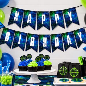 Laser tag birthday banner is displayed above a colorful party table with bright blue, neon green and black decorations. The 5x7" pennants spell Happy Birthday in bold distressed white lettering over glowing blue and green laser graphics. You've Been Tagged end pieces feature coordinating neon target designs. Balloons, cupcakes and gamer party decorations complete the laser tag birthday setup.