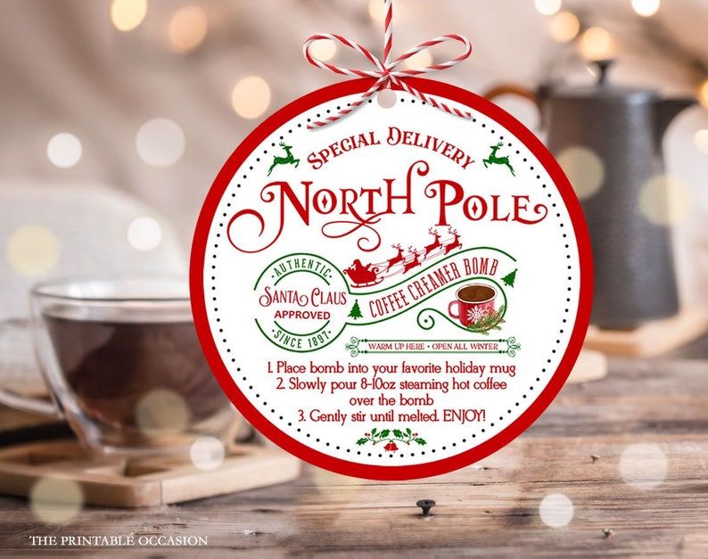 May include: A red and white circular tag with the words "North Pole" and "Coffee Creamer Bomb." The tag features a Santa Claus graphic and instructions for use. A clear glass mug with a dark liquid is in the foreground.